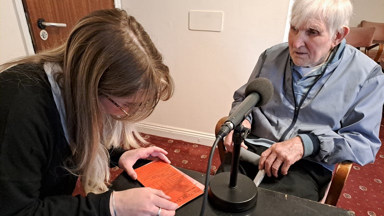 A microphone sat on a table facing and elderly man, with a female looking at City of Bath rent books.